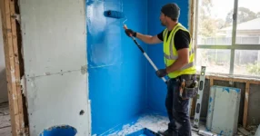 Licensed waterproofer rolling bright blue waterproofing membrane onto a shower wall in an unfinished Australian bathroom renovation